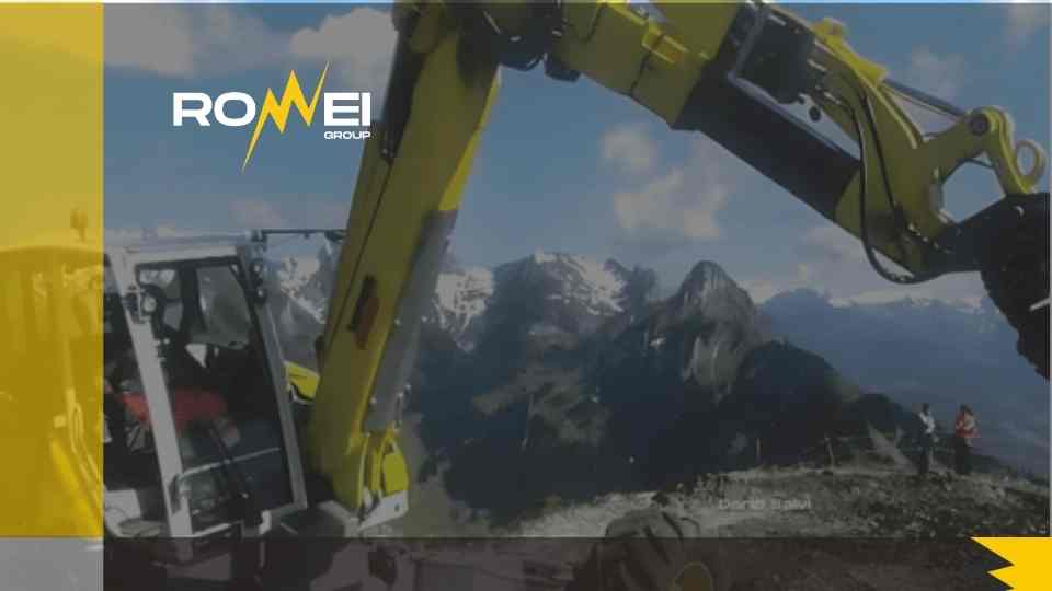 Un'inquadratura dall'alto del braccio esteso di un escavatore giallo in un cantiere di montagna con una persona che lo aziona nella cabina. Il logo "ROMEI GROUP" si trova nell'angolo in alto a sinistra.