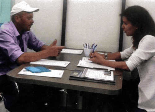 Un uomo con una maglietta lilla e un cappellino bianco è seduto a un tavolo durante un'intervista. Di fronte a lui, una donna prende appunti su alcuni fogli. Al centro del tavolo è presente un portapenne.