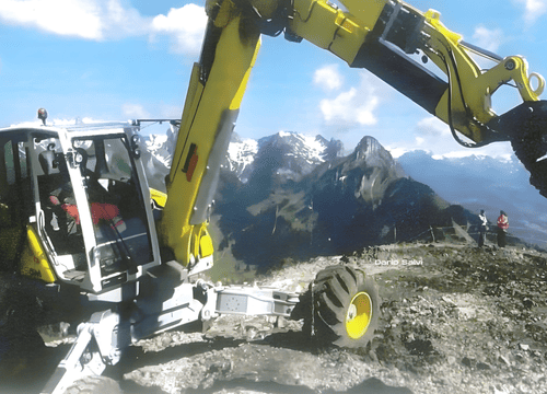 Primo piano di un escavatore ragno giallo e grigio posizionato su un terreno roccioso in alta montagna. Il braccio meccanico è sollevato verso l'alto, con uno sfondo di vette alpine innevate sotto un cielo azzurro.