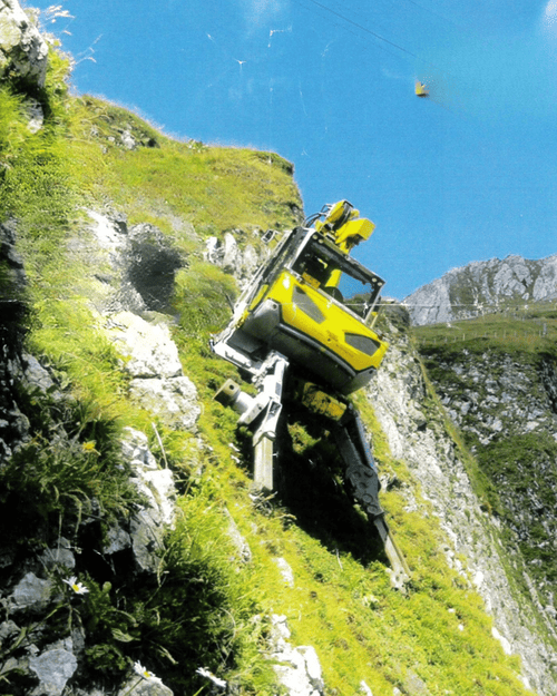 Un escavatore ragno giallo opera in condizioni estreme su un pendio erboso e roccioso molto scosceso. La macchina è inclinata e utilizza le sue zampe articolate per stabilizzarsi sulla parete della montagna.